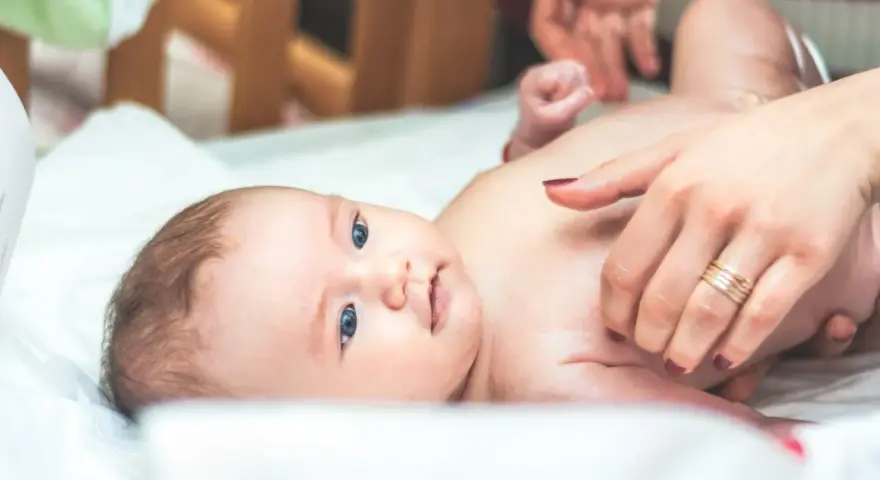 Niebieskooki bobas leży na białym prześcieradle, a jego mama delikatnie go dotyka. Na palcu mamy widać obrączkę, a na jej paznokciach ciemny lakier. Dziecko ma na sobie tylko pieluszkę, a jego skóra jest gładka i delikatna, jak **perły Epsteina**.