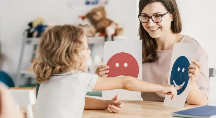 Dziecko i pedagog pracują nad emocjami podczas terapii pedagogicznej, używając kart z buźkami.