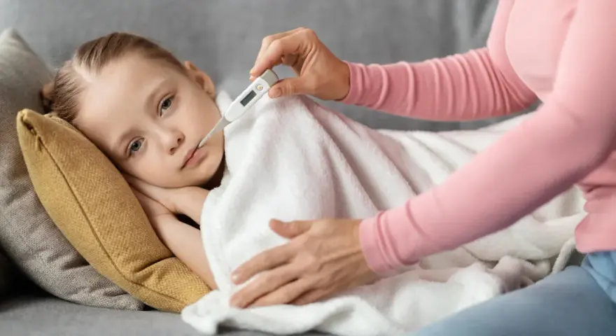 Dziecko leży pod kocem, a mama mierzy mu temperaturę. Wysoka gorączka u dziecka bez innych objawów martwi.