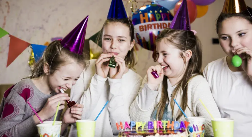 Radosne zabawy na urodziny! Dziewczynki w stożkowych czapkach dmuchają w gwizdki, śmiejąc się przy torcie z napisem "Happy Birthday".