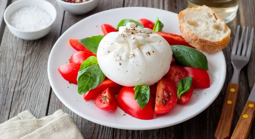 Burrata z pomidorami i bazylią na talerzu, obok pieczywo i przyprawy.