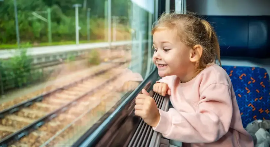 Uśmiechnięta dziewczynka patrzy przez okno pociągu. Zastanawia się, od ilu lat można jeździć samemu pociągiem, marząc o podróżach.