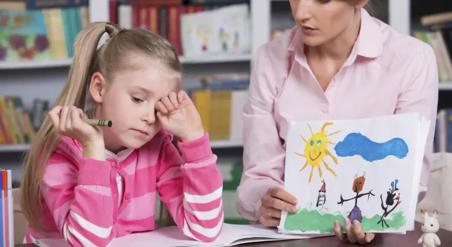 Dziewczynka z kredką w dłoni, smutna, podczas sesji socjoterapii. Terapeuta pokazuje jej rysunek.