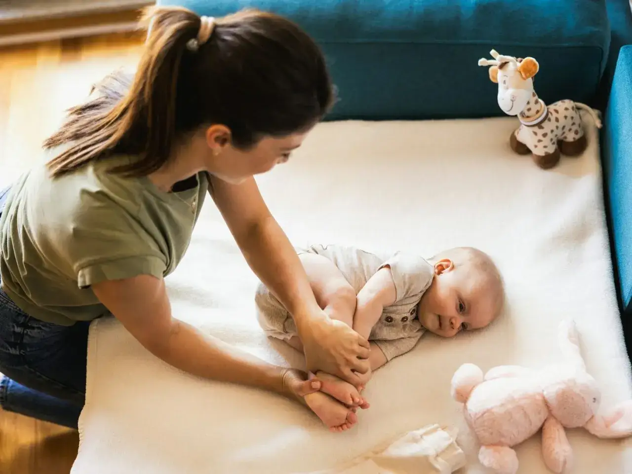 Mama bawi się z 6-miesięcznym dzieckiem na białym kocyku. Obok leżą pluszowe zabawki: żyrafa i króliczek.