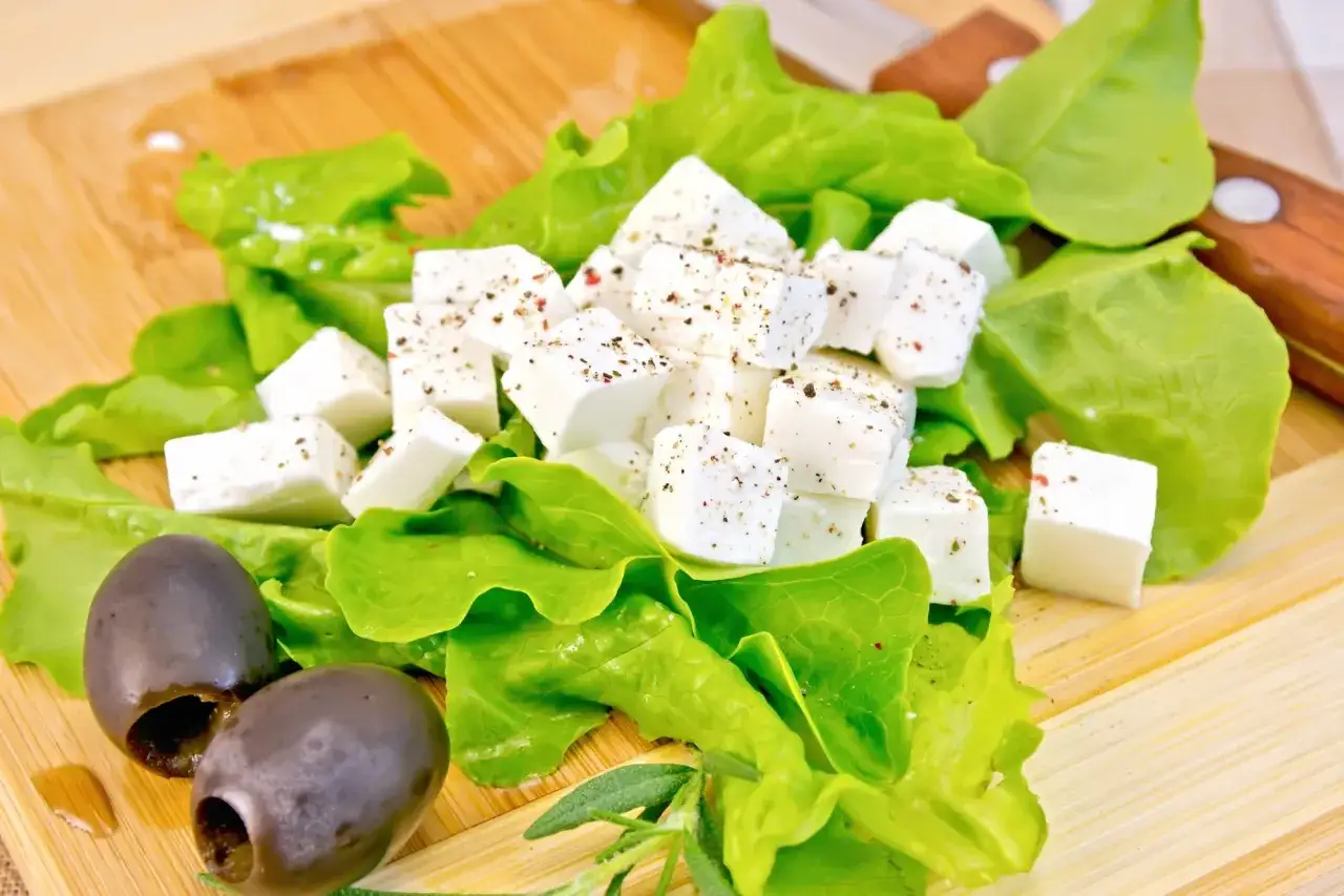 Kostki sera feta w ciąży, posypane ziołami, na liściach sałaty z oliwkami.