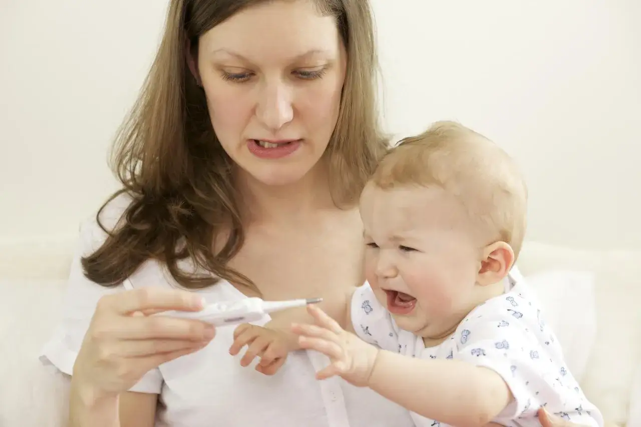 Zmartwiona mama mierzy temperaturę płaczącemu dziecku, które ma gorączkę po szczepieniu.