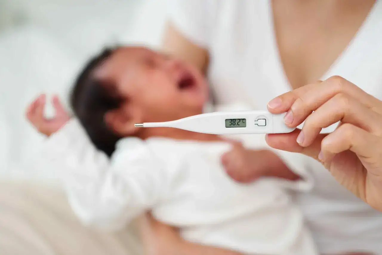 Termometr pokazuje 38.2°C. Mama sprawdza prawidłową temperaturę u niemowlaka na czole, martwiąc się o jego zdrowie.