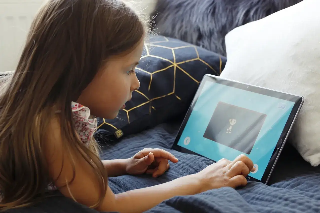 Mała dziewczynka, typowy ipad kid, leży na łóżku i z zaciekawieniem ogląda bajkę na tablecie.