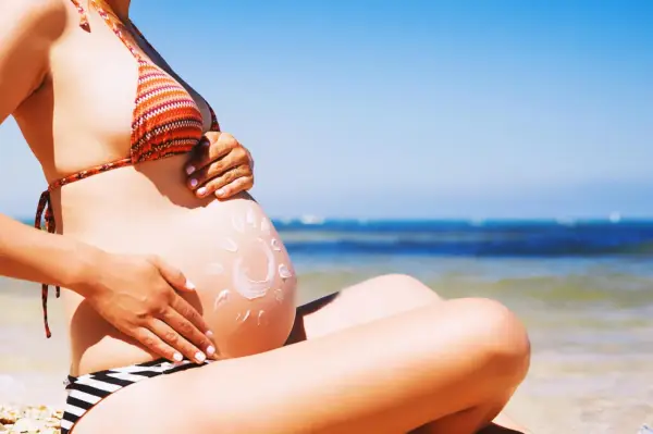 Ciężarna kobieta na plaży, z kremem przeciwsłonecznym na brzuchu w kształcie słońca. Bezpieczne opalanie natryskowe w ciąży.